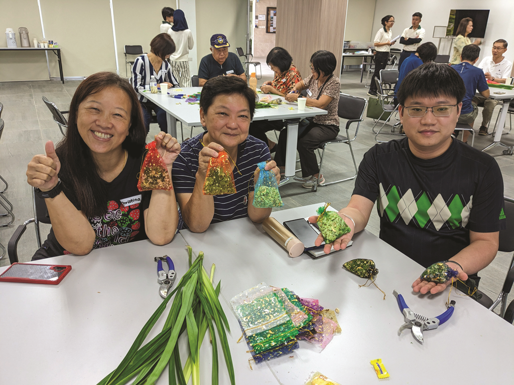A group of people are sitting around tables making sachets filled with aromatic herbs. They are holding up the finished sachets and smiling.